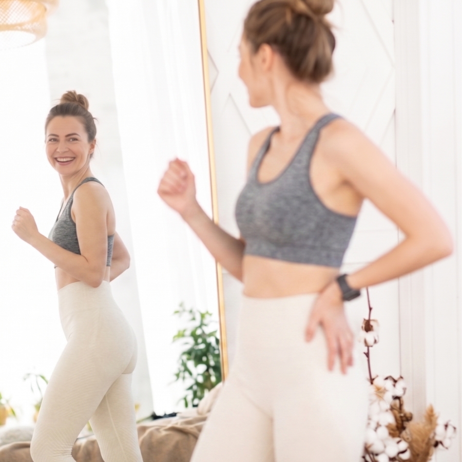 Woman in gray sports bra and light leggings looks in mirror and smiles during a workout in a bright room.