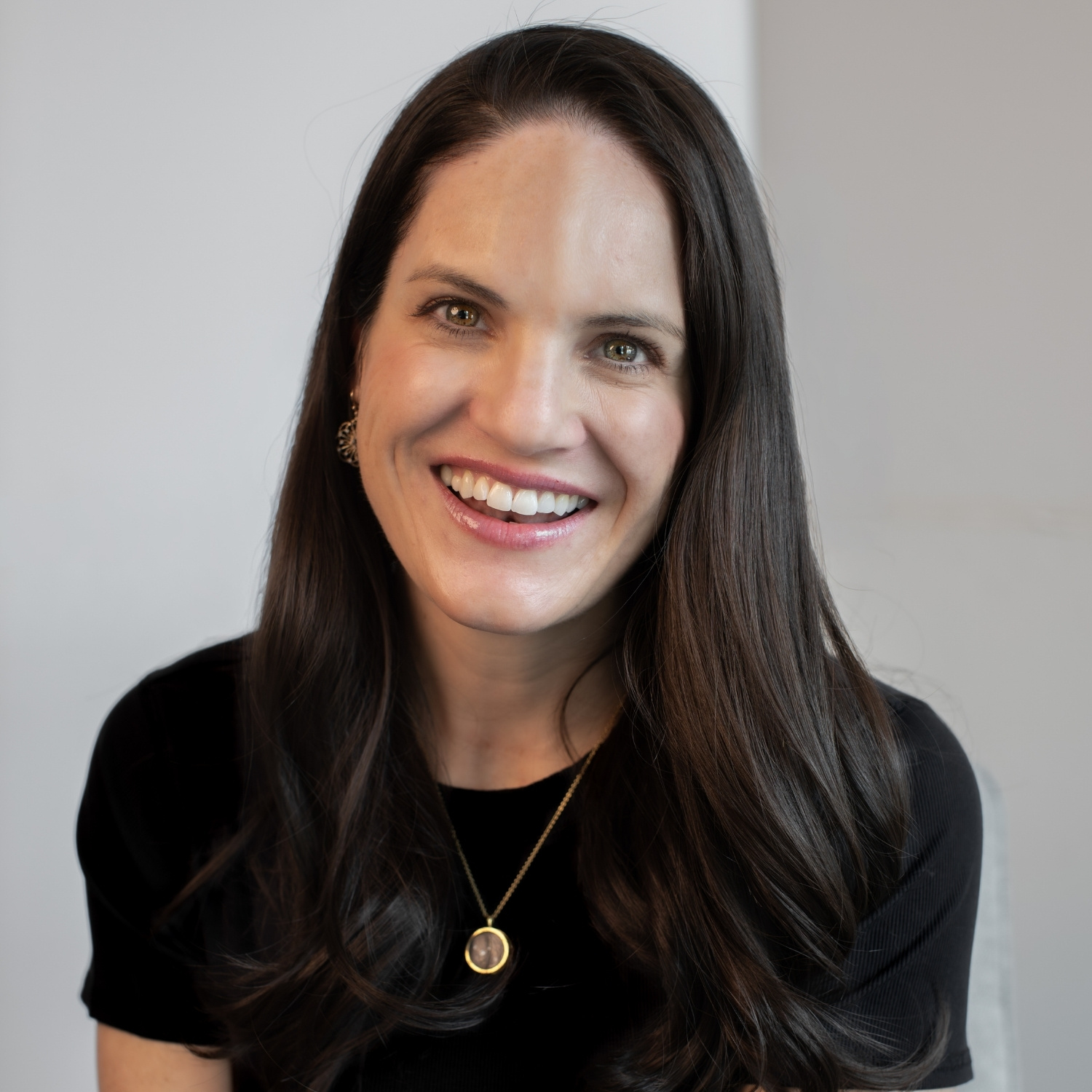 Portrait of a smiling woman with long dark hair wearing a black top and a gold pendant necklace against a neutral background.