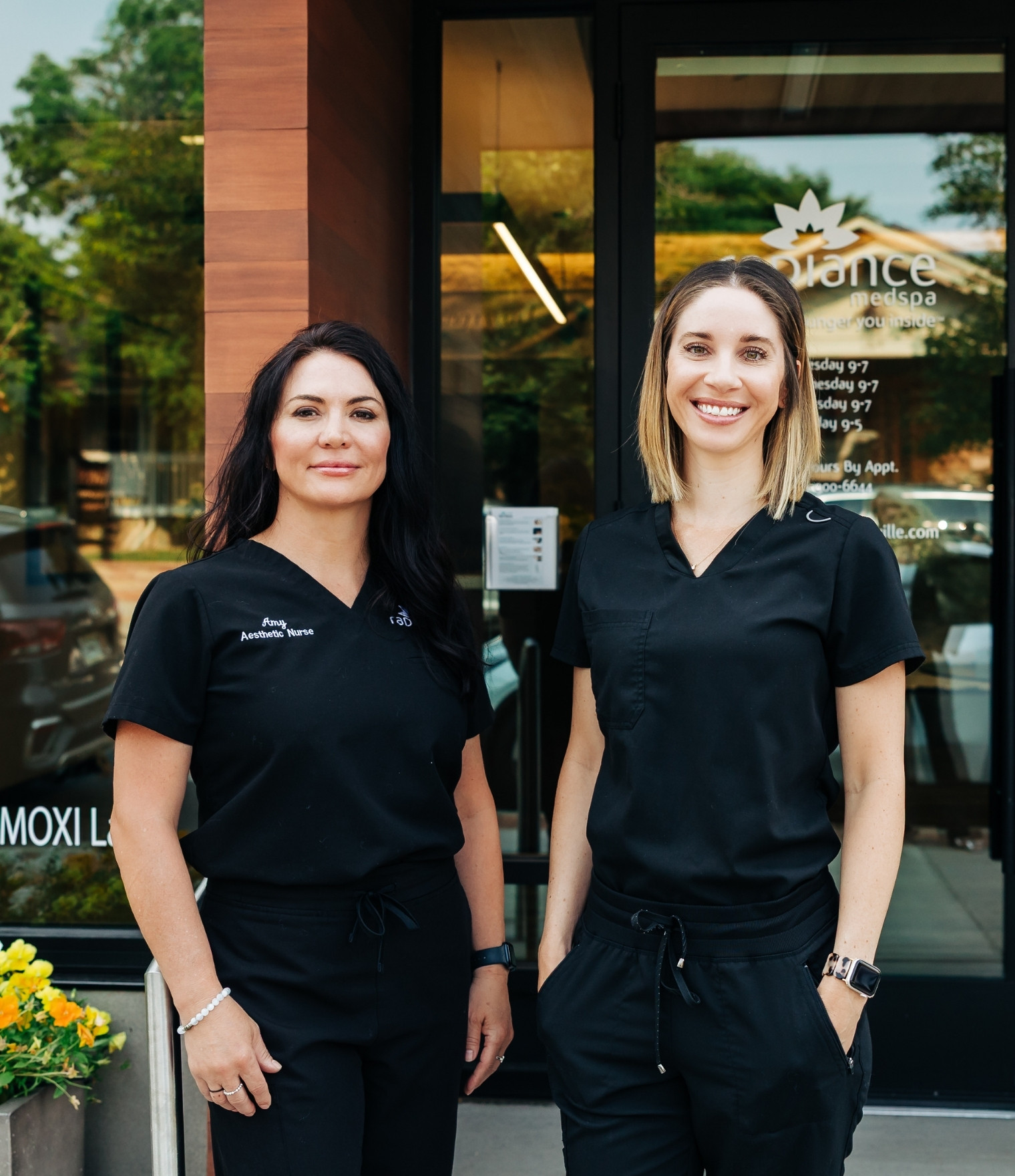 Two smiling women in black scrubs stand outside a spa or clinic storefront, posing for the photo.