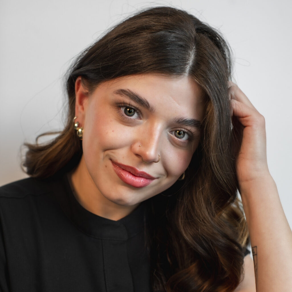 Close-up of a smiling woman with long dark hair, nose ring, and gold earrings, touching her hair.